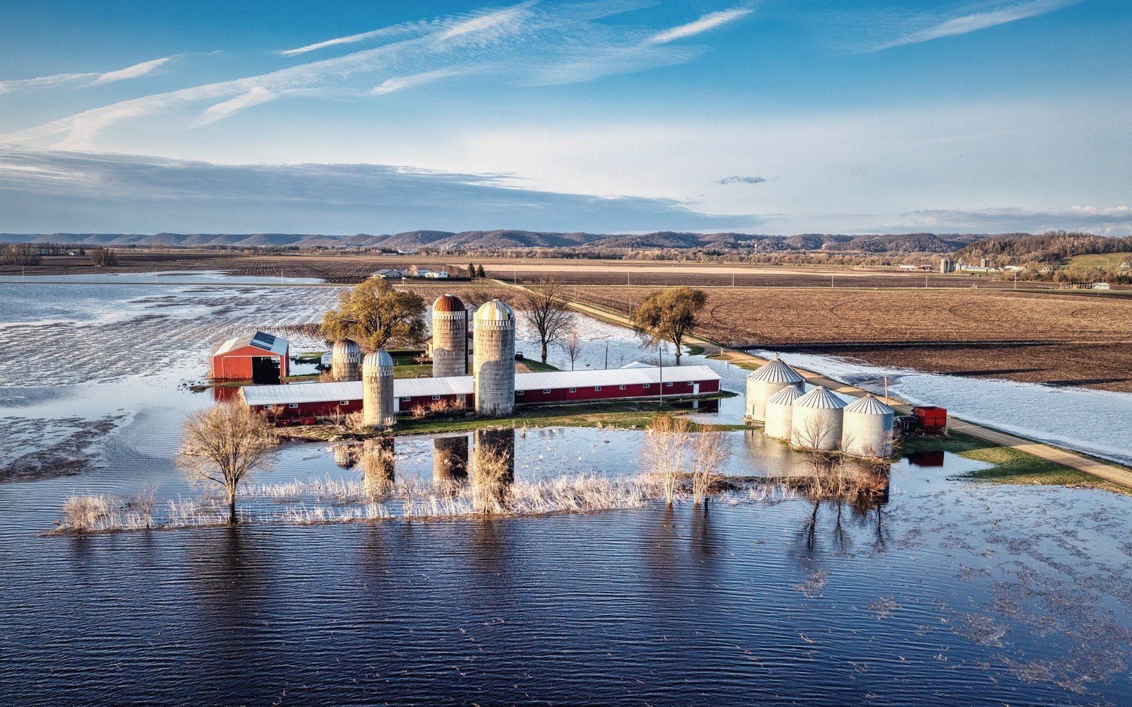 Aerial view of a flooded farm with silos and barns surrounded by rising floodwater