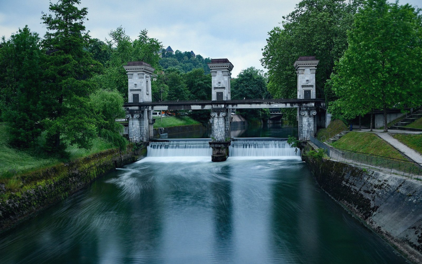 Water flows over a river dam with three stone towers connecting a footbridge, surrounded by trees