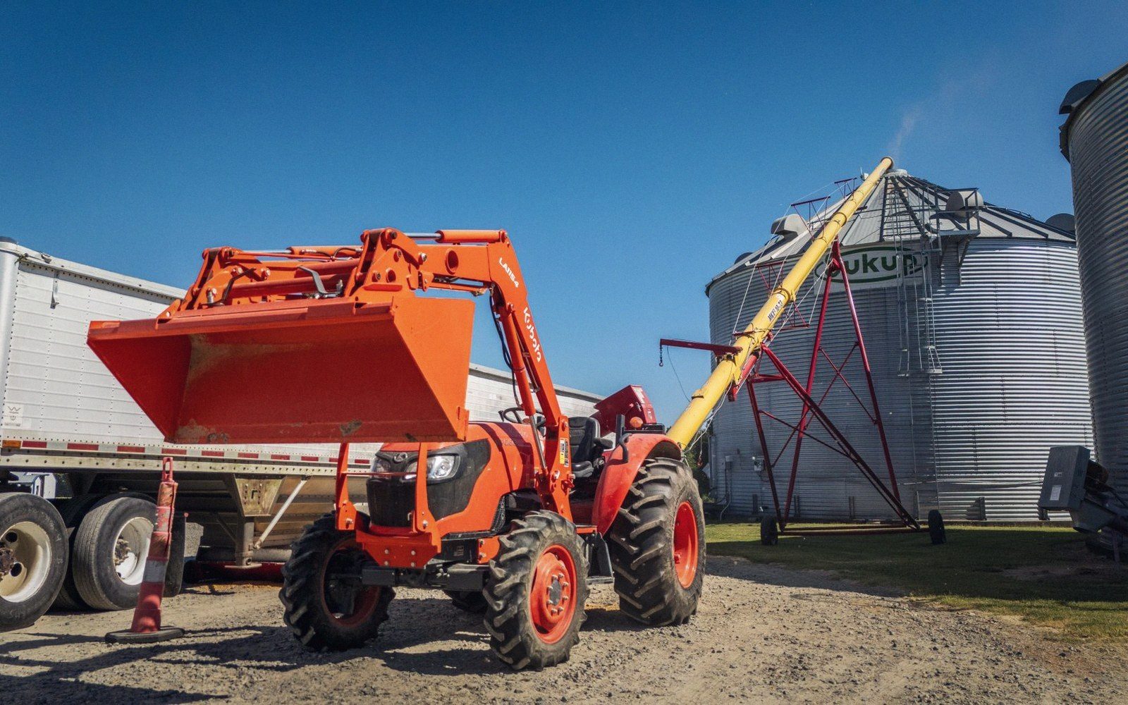 Orange tractor with front loader parked near grain bins and an auger