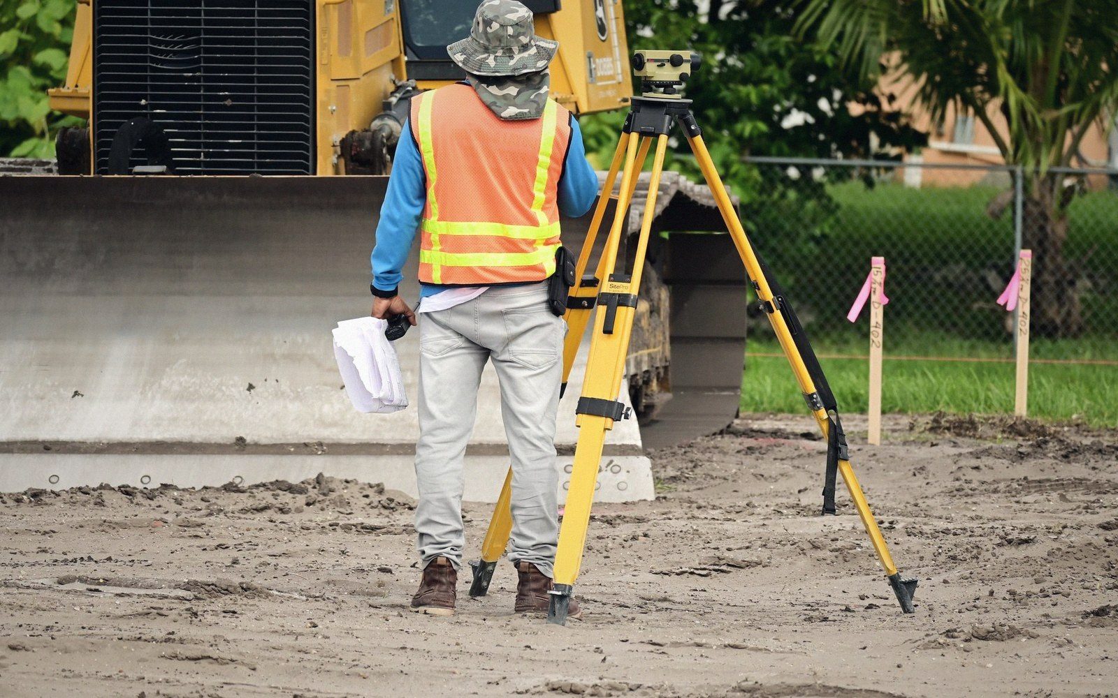 Surveyor in safety vest stands beside a tripod-mounted level near a bulldozer on a construction site